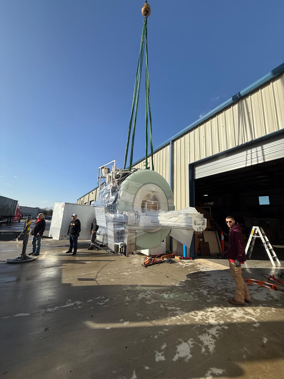 Crane lifting a shrink-wrapped Siemens MRI magnet outside a warehouse while the rigging crew looks on
